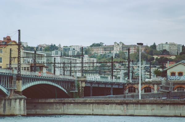Un pont ferroviaire au-dessus du Rhône, avec beaucoup de poteaux et de câbles, on voit les hauteurs de la ville en arrière-plan en arrière plan avec un joli bâtiment XIXième.